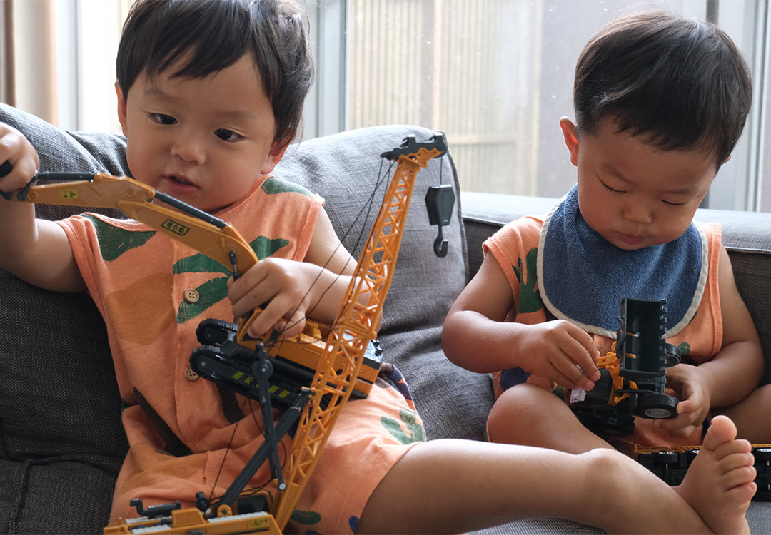 双子の男の子が車で遊んでいる。それぞれクレーン車とトラクターを手に持っている。