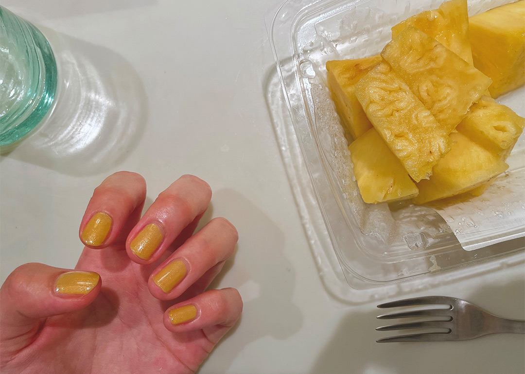 OSAJIの黄色のネイルをするKanocoさんの手と食べやすいようにカットされパックに入ったままのパイナップルが並んでいる。
